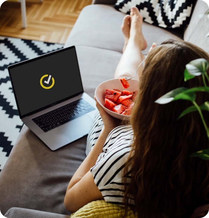 Nainen syö vesimelonia sohvalla käyttäen kannettavaa tietokonetta, jossa käynnistyy Norton 360 AntiVirus Plus